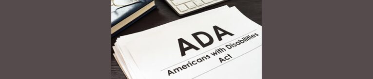 A stack of papers titled "ADA Americans with Disabilities Act" sits on a desk next to a keyboard, a book, and a guide on "How to Check Your Website for ADA Compliance.