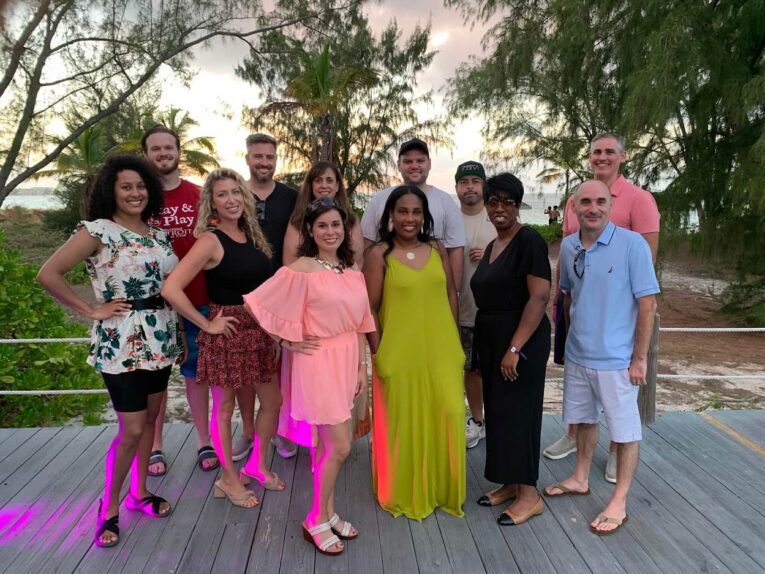 A group of twelve people posing on a wooden deck with trees and a beach in the background. Some are standing while others have one foot forward or hands on hips. They are dressed in casual and semi-formal attire.